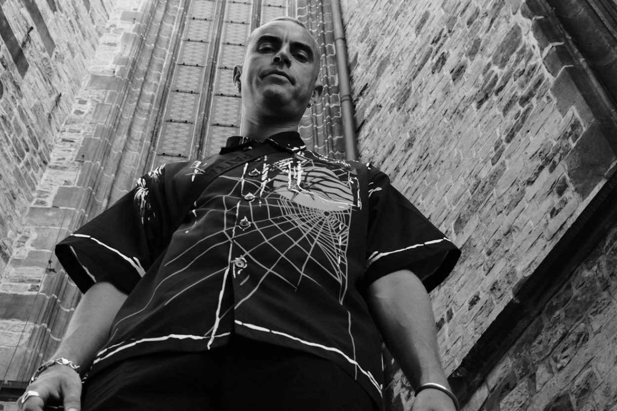 Alix Perez in un ritratto dal basso davanti a un'architettura gotica, camicia a ragnatela