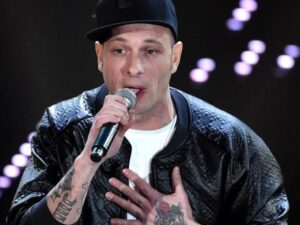 Clementino durante un'esibizione live al microfono, con giacca in pelle e tatuaggi in primo piano