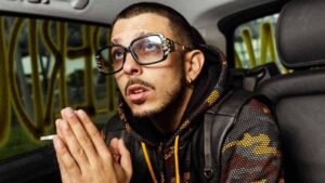 Enzo Dong seduto sul sedile posteriore di un'auto con occhiali vistosi e sigaretta in mano