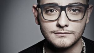 Ritratto ravvicinato di Rocco Hunt con occhiali da vista e catena d'argento al collo