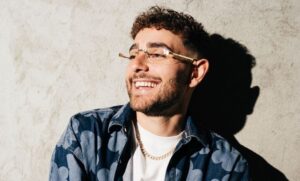 Rocco Hunt sorridente con capelli ricci e occhiali da sole al collo, ritratto informale