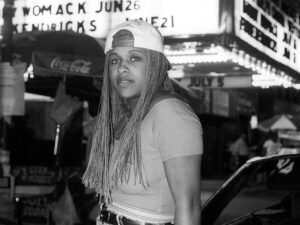 Ritratto in bianco e nero della rapper Yo-Yo con treccine e cappellino, di sera davanti a un cinema