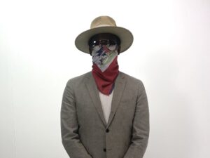Mach-Hommy con cappello a tesa larga, occhiali, bandana rossa sul volto e giacca grigia su sfondo bianco
