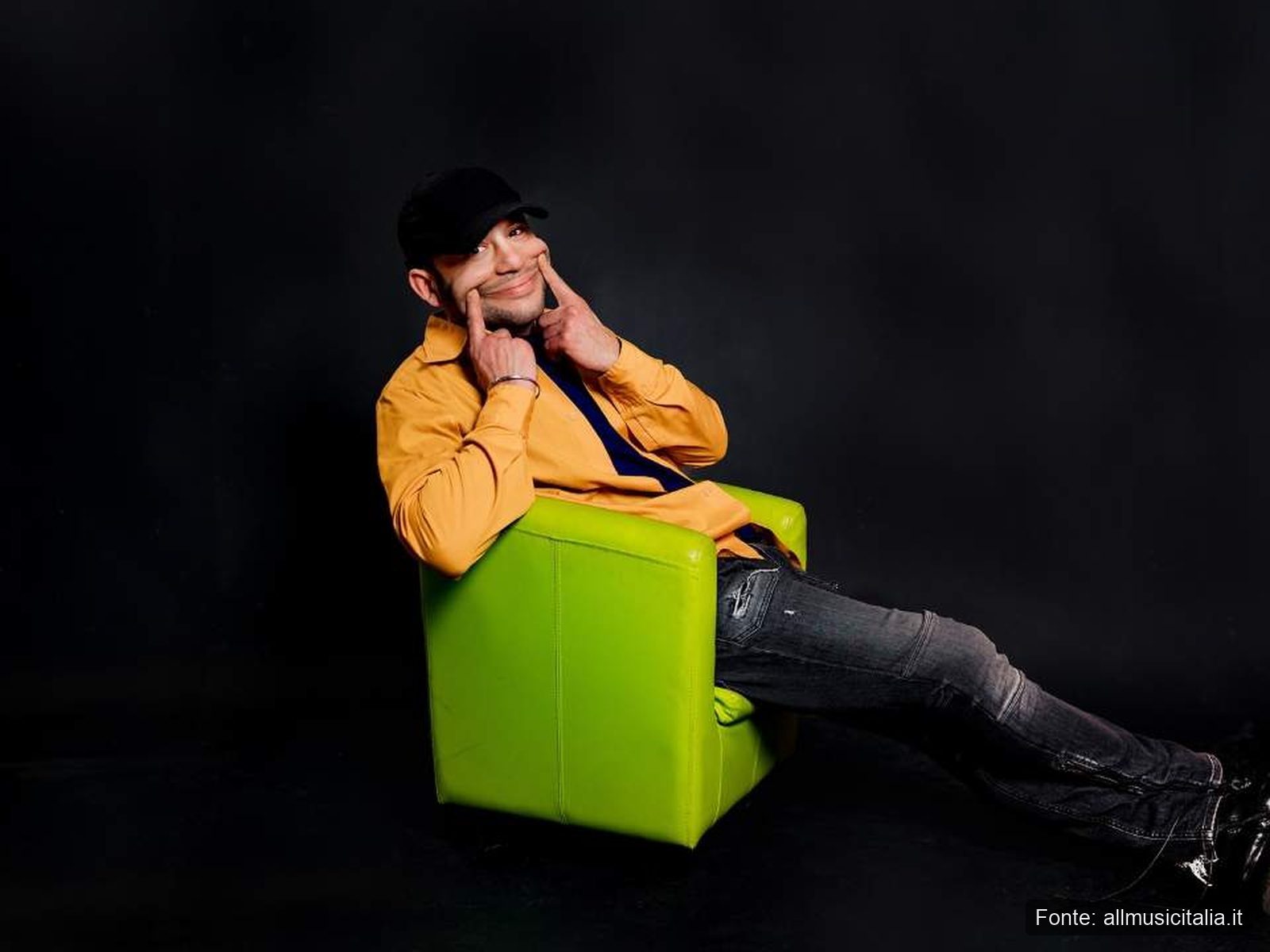 Rancore Dj Myke Sunshine Foto Poltrona Verde 2026 Rancore seduto su una poltrona verde mentre si stira il sorriso con le dita
