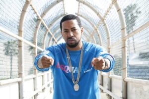Roc Marciano con felpa blu e catena su una passerella coperta con rete metallica, pugni verso la camera