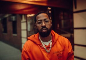 Roc Marciano in ritratto con occhiali e catena d'argento, felpa arancione, in un ambiente illuminato di sera