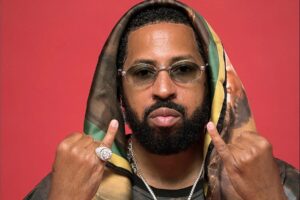 Roc Marciano con occhiali, anello e foulard sul capo davanti a uno sfondo rosso, mani in segno di gesto