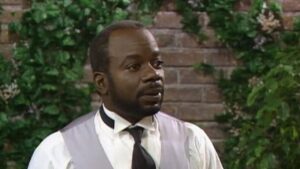 Joseph Marcell nel ruolo del maggiordomo Geoffrey con panciotto grigio e cravatta, davanti a un muro di edera in Willy, il principe di Bel-Air
