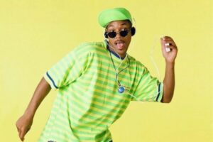 Will Smith con cuffie, occhiali scuri e cappellino verde in una maglia a righe, in posa su sfondo giallo per Willy, il principe di Bel-Air