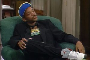 Will Smith con cappellino blu, giacca nera e sneakers Jordan sprofondato in una poltrona verde in una scena di Willy, il principe di Bel-Air